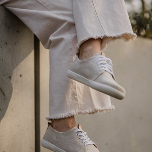May include: A pair of off-white canvas sneakers with white laces. The sneakers are worn by a person wearing light-colored pants.