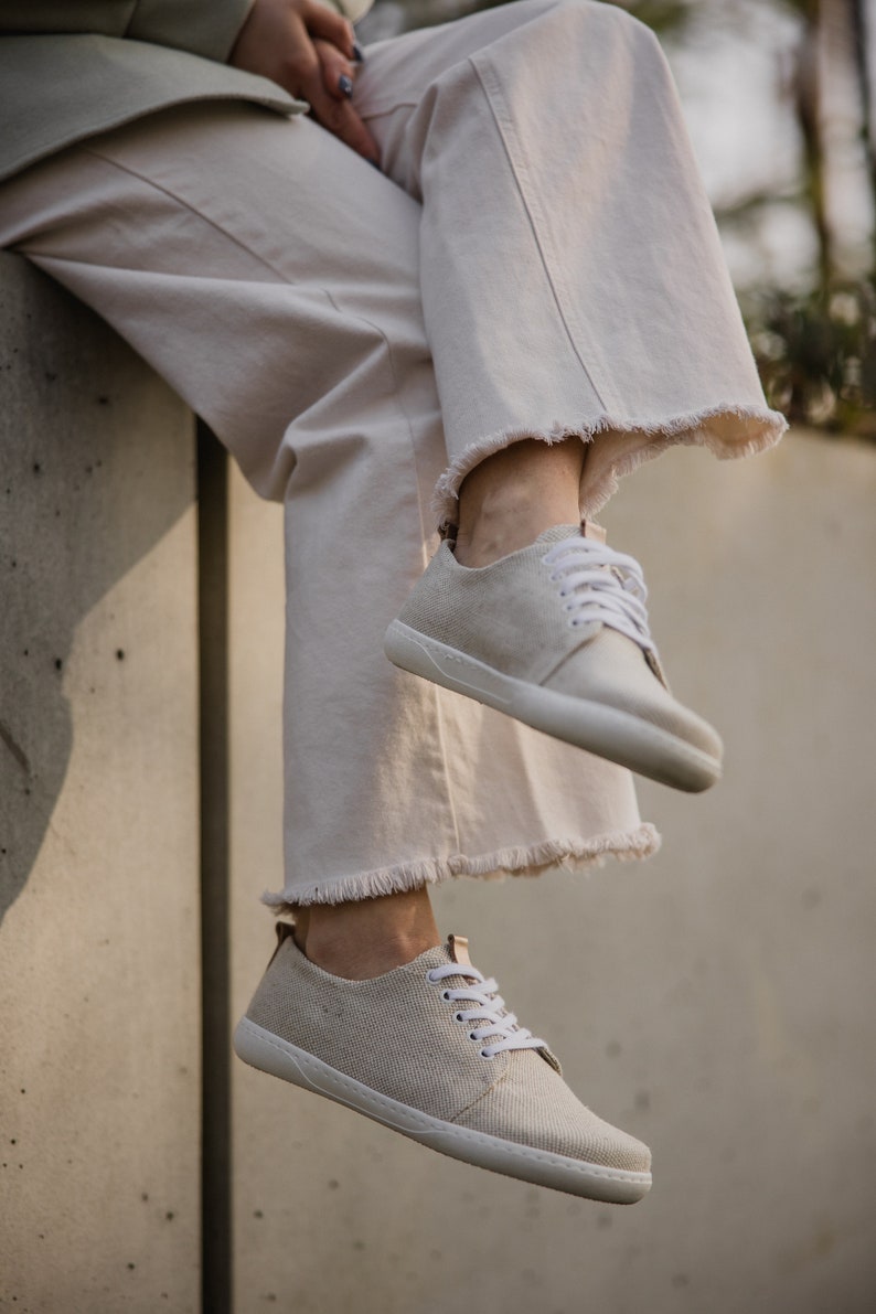 May include: A pair of off-white canvas sneakers with white laces. The sneakers are worn by a person wearing light-colored pants.