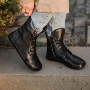 Könnte beinhalten: Schwarze Leder-Ankle Boots mit Schnürung vorne und seitlichem Reißverschluss. Die Stiefel haben eine runde Spitze und eine flache Sohle. Getragen mit blauen Jeans.