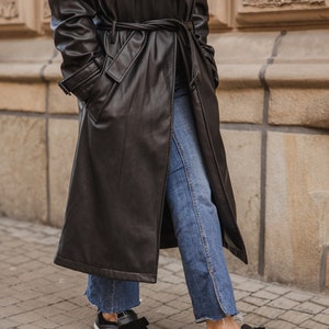 Pode incluir: Uma pessoa vestindo um longo casaco de couro preto com cinto, jeans azul e sapatos pretos com detalhe de la&ccedil;o.