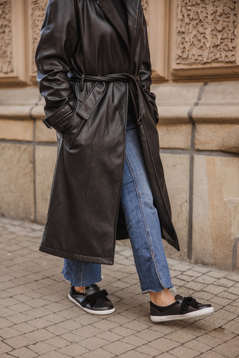 Pode incluir: Uma pessoa vestindo um longo casaco de couro preto com cinto, jeans azul e sapatos pretos com detalhe de la&ccedil;o.