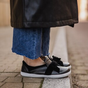 Pode incluir: Sapatos pretos de couro com um grande la&ccedil;o preto na parte superior. Os sapatos t&ecirc;m uma sola branca e s&atilde;o usados por uma pessoa em p&eacute; em uma cal&ccedil;ada de tijolos.
