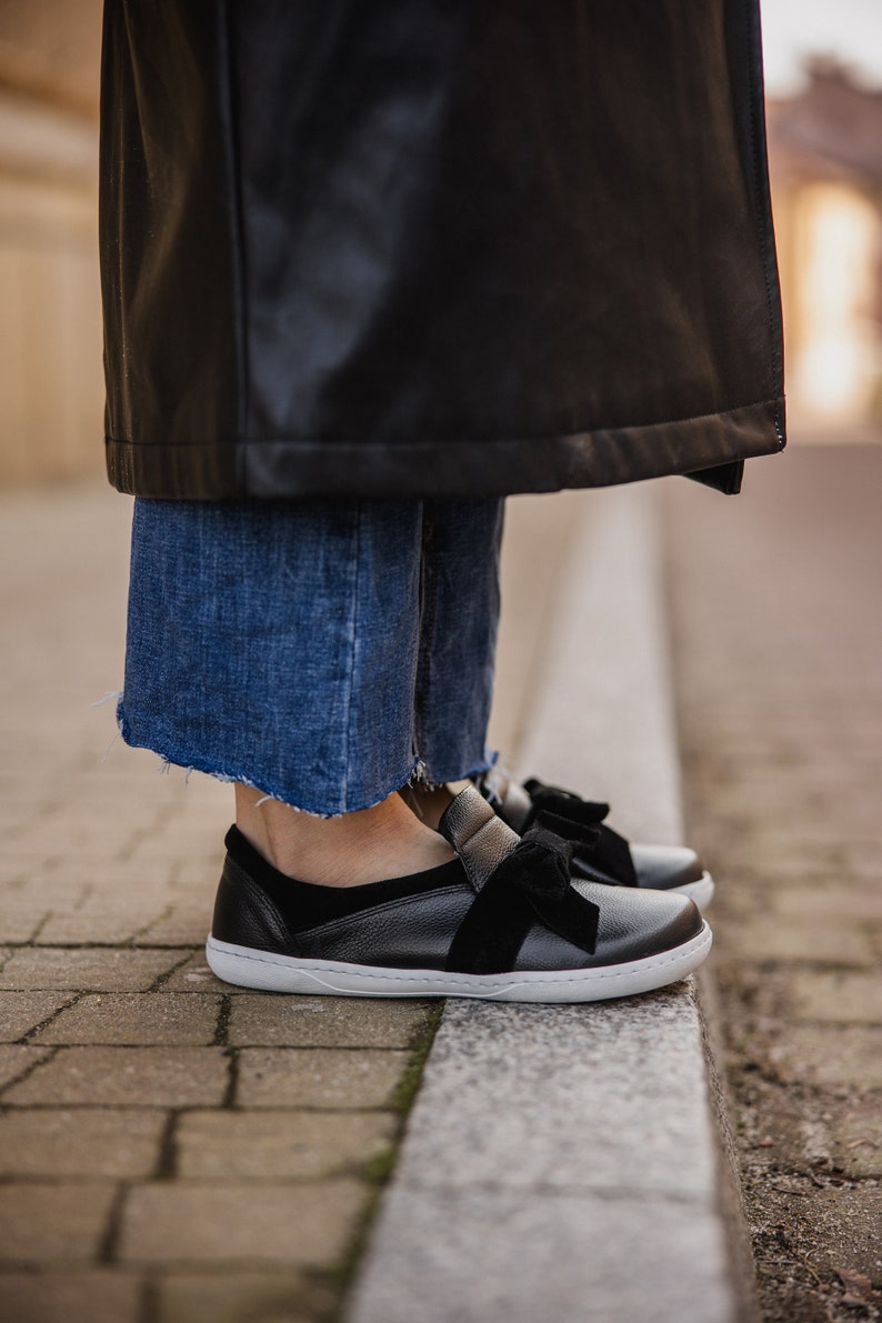 Pode incluir: Sapatos pretos de couro com um grande la&ccedil;o preto na parte superior. Os sapatos t&ecirc;m uma sola branca e s&atilde;o usados por uma pessoa em p&eacute; em uma cal&ccedil;ada de tijolos.