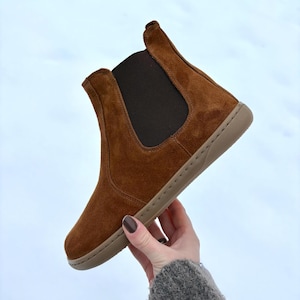 May include: A brown suede Chelsea boot with a tan sole and elastic side panel. The ankle boot has a rounded toe and a pull tab at the back. The boot is held up against a white background.