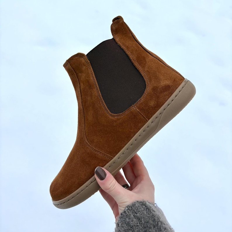 May include: A brown suede Chelsea boot with a tan sole and elastic side panel. The ankle boot has a rounded toe and a pull tab at the back. The boot is held up against a white background.