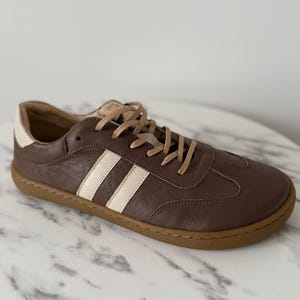 Op de afbeelding: Bruine leren sneakers met cr&egrave;mekleurige strepen en een bruine zool. De schoenen hebben beige veters en een witte hielkap. Deze casual sneakers zijn geschikt voor dagelijks gebruik.
