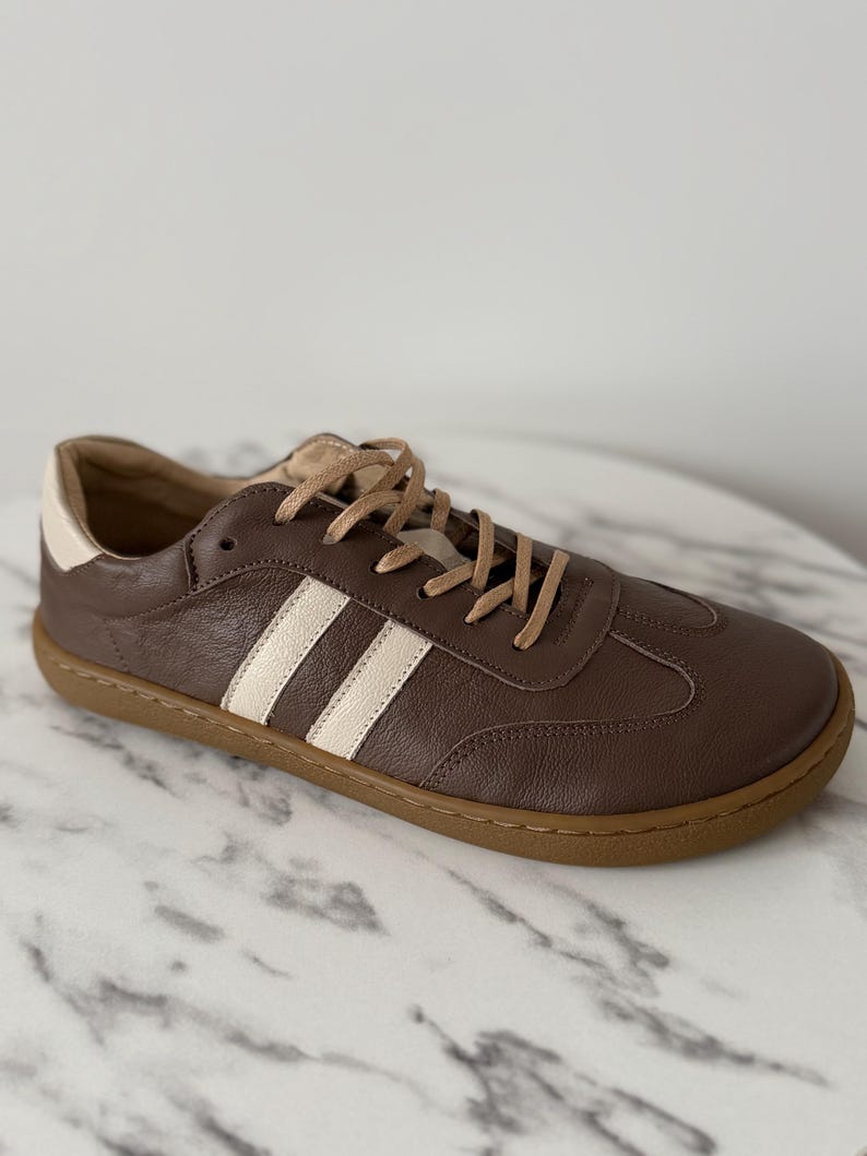 Op de afbeelding: Bruine leren sneakers met cr&egrave;mekleurige strepen en een bruine zool. De schoenen hebben beige veters en een witte hielkap. Deze casual sneakers zijn geschikt voor dagelijks gebruik.