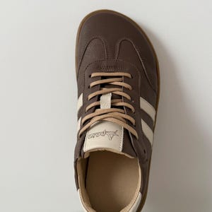 Op de afbeelding: Bruine leren sneaker met cr&egrave;mekleurige strepen en veters. De schoen heeft een ronde neus en een lichtbruine zool. De merknaam "Apareo" is zichtbaar op de tong. De sneaker is ontworpen voor casual wear.