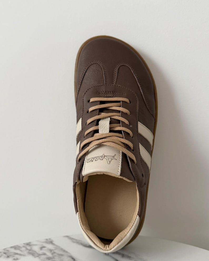 Op de afbeelding: Bruine leren sneaker met cr&egrave;mekleurige strepen en veters. De schoen heeft een ronde neus en een lichtbruine zool. De merknaam "Apareo" is zichtbaar op de tong. De sneaker is ontworpen voor casual wear.