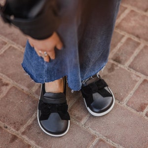 Pode incluir: Sapatos pretos de couro com um la&ccedil;o de veludo preto na frente. Os sapatos t&ecirc;m uma sola de borracha branca.
