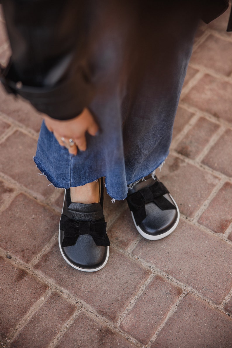 Pode incluir: Sapatos pretos de couro com um la&ccedil;o de veludo preto na frente. Os sapatos t&ecirc;m uma sola de borracha branca.