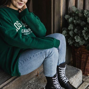 Op de afbeelding: Een vrouw draagt een groen sweatshirt met de tekst "LOS G. CALORN Long Beach, CA" en zit met gekruiste benen op een houten trede. Ze draagt een blauwe jeans en zwarte veterschoenen.