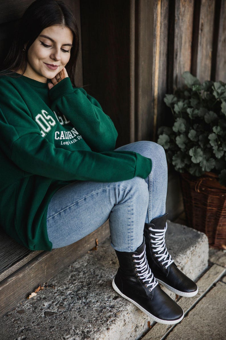 Op de afbeelding: Een vrouw draagt een groen sweatshirt met de tekst "LOS G. CALORN Long Beach, CA" en zit met gekruiste benen op een houten trede. Ze draagt een blauwe jeans en zwarte veterschoenen.