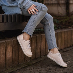 May include: A pair of light pink leather sneakers with white laces. The sneakers are worn by a person sitting on a bench.