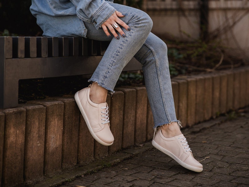 May include: A pair of light pink leather sneakers with white laces. The sneakers are worn by a person sitting on a bench.