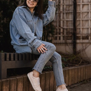 May include: A woman wearing a blue denim jacket and jeans sits on a bench wearing light pink sneakers. She is smiling and wearing round sunglasses.