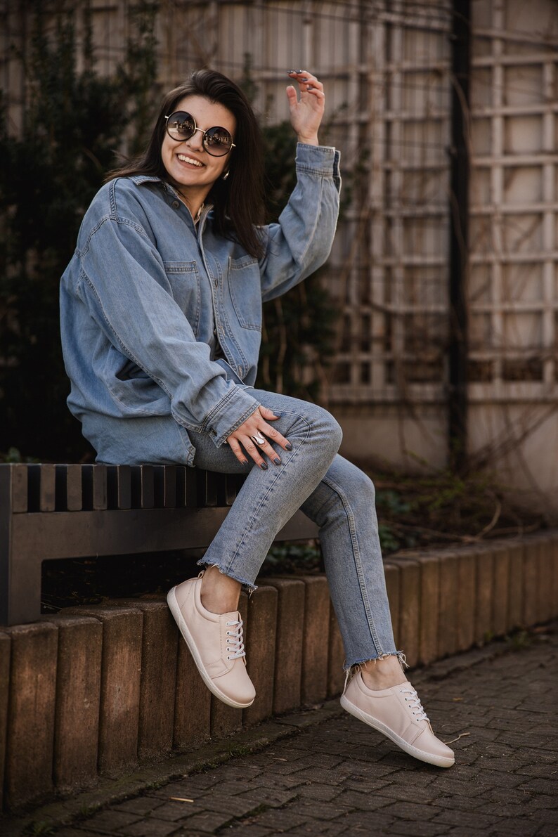 May include: A woman wearing a blue denim jacket and jeans sits on a bench wearing light pink sneakers. She is smiling and wearing round sunglasses.