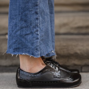 Pu&ograve; includere: Scarpe Oxford in pelle nera con perforazioni decorative e finitura lucida. Sono abbinate a jeans blu con orlo sfilacciato, creando un look casual. Le scarpe sono su una superficie di cemento.