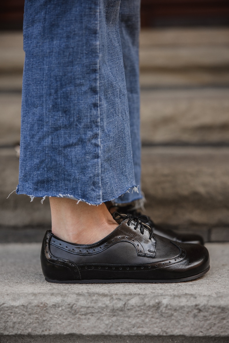Pu&ograve; includere: Scarpe Oxford in pelle nera con perforazioni decorative e finitura lucida. Sono abbinate a jeans blu con orlo sfilacciato, creando un look casual. Le scarpe sono su una superficie di cemento.