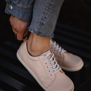 May include: A pair of light pink leather sneakers with white laces. The sneakers are worn by a person wearing blue jeans.