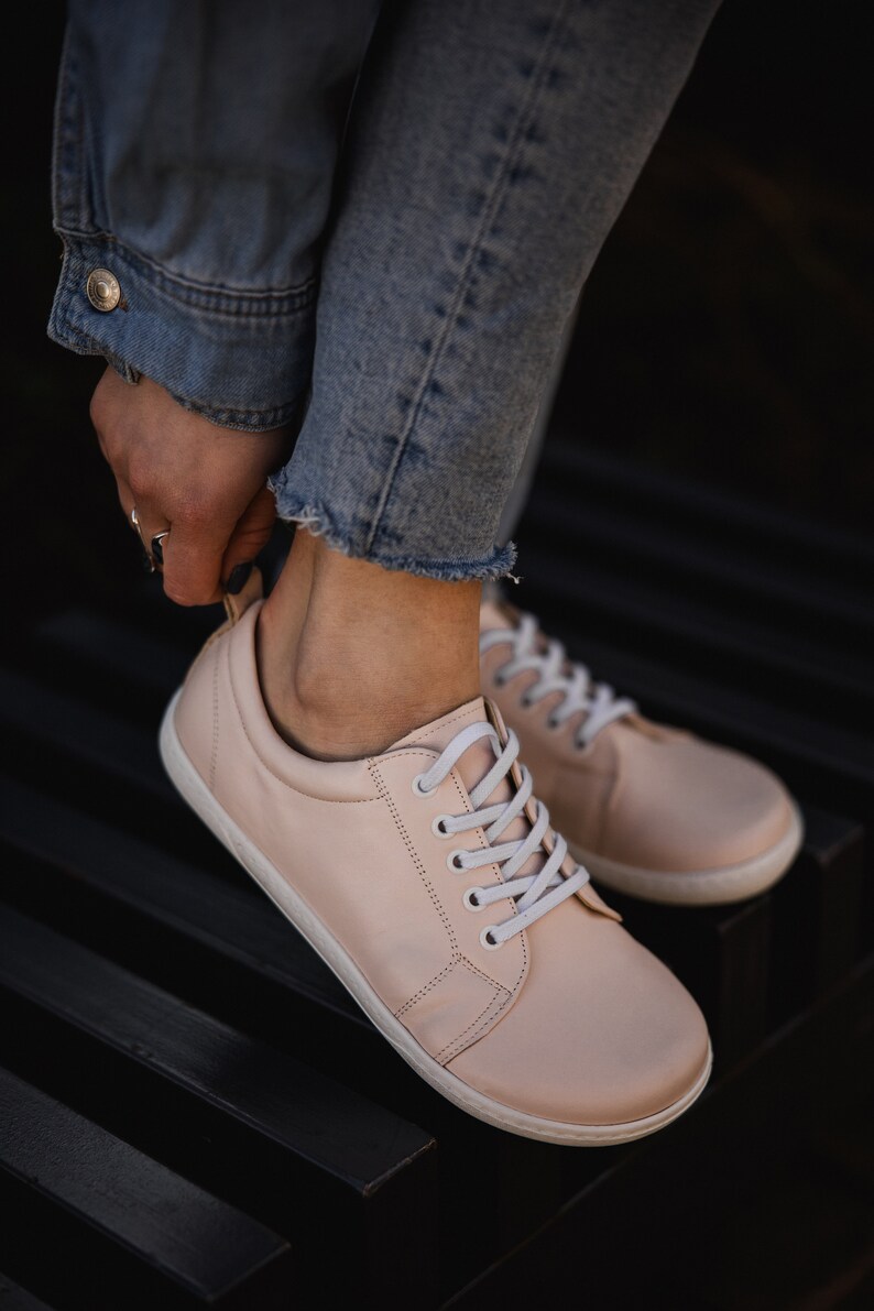 May include: A pair of light pink leather sneakers with white laces. The sneakers are worn by a person wearing blue jeans.