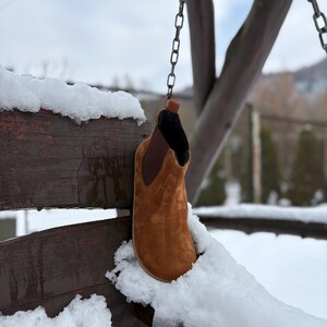 May include: A brown suede ankle boot hangs from a chain in a snowy outdoor scene. The boot has a rounded toe and a dark brown elastic side panel. The background features a wooden structure and a snow-covered landscape.