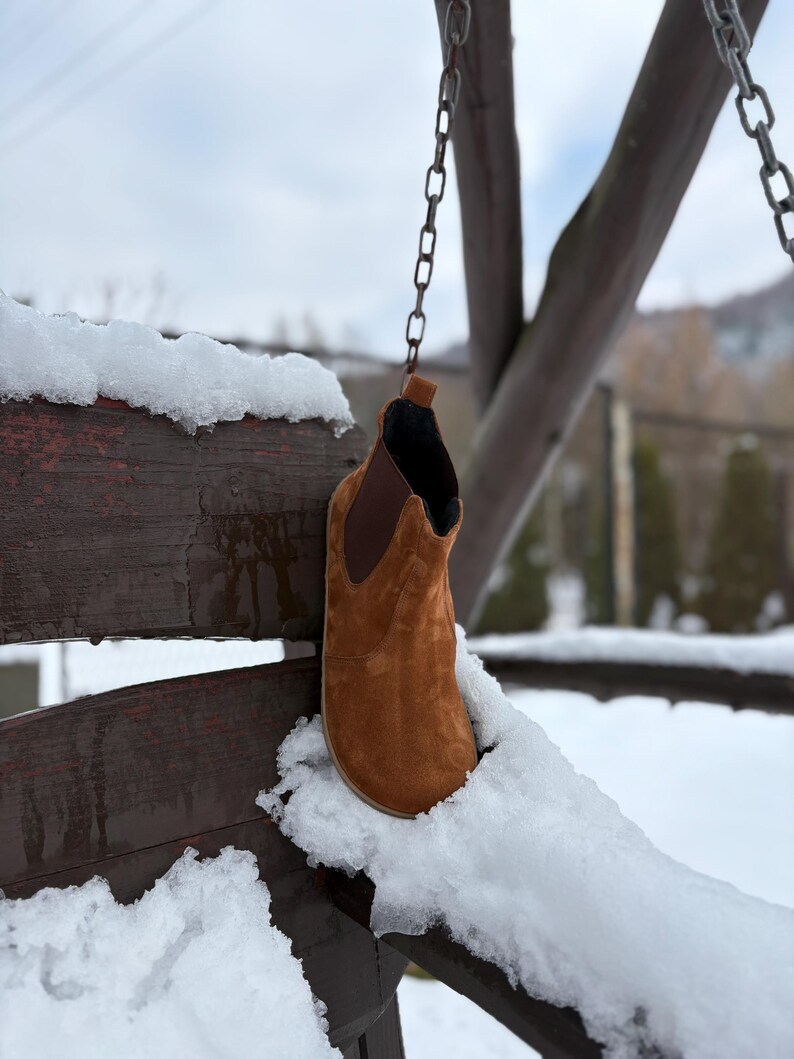 May include: A brown suede ankle boot hangs from a chain in a snowy outdoor scene. The boot has a rounded toe and a dark brown elastic side panel. The background features a wooden structure and a snow-covered landscape.