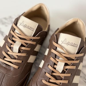 Op de afbeelding: Bruine leren sneakers met cr&egrave;mekleurige strepen en veters. De tong van elke schoen heeft de merknaam "Apavo". De sneakers hebben een klassiek, sportief ontwerp.