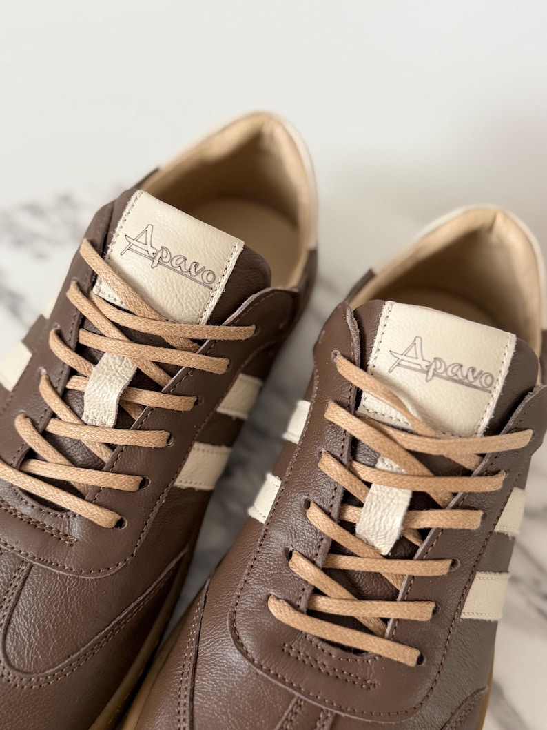 Op de afbeelding: Bruine leren sneakers met cr&egrave;mekleurige strepen en veters. De tong van elke schoen heeft de merknaam "Apavo". De sneakers hebben een klassiek, sportief ontwerp.