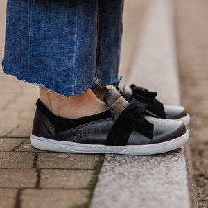Pode incluir: Sapatos pretos de couro com um detalhe de la&ccedil;o de veludo preto. Os sapatos t&ecirc;m uma sola de borracha branca e s&atilde;o usados com cal&ccedil;as de ganga azuis.