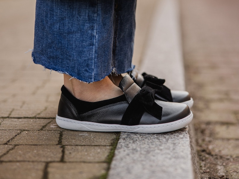 Pode incluir: Sapatos pretos de couro com um detalhe de la&ccedil;o de veludo preto. Os sapatos t&ecirc;m uma sola de borracha branca e s&atilde;o usados com cal&ccedil;as de ganga azuis.