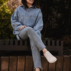 May include: A woman wearing a light blue denim shirt and jeans sits on a black bench. She is wearing white sneakers and sunglasses.