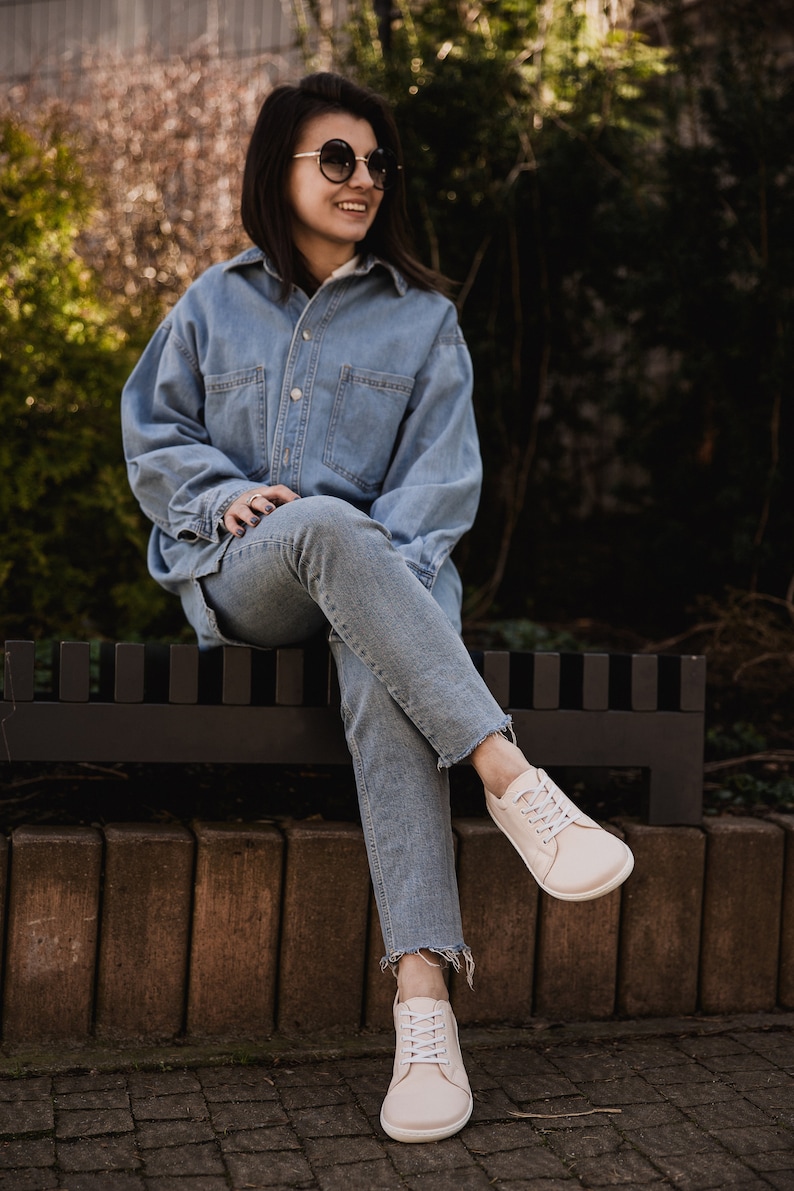May include: A woman wearing a light blue denim shirt and jeans sits on a black bench. She is wearing white sneakers and sunglasses.