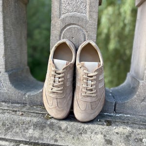 Può includere: Un paio di sneakers in camoscio beige con lacci e dettagli color crema. Le scarpe hanno un design classico e sono adatte per l'abbigliamento casual. Le sneakers sono esposte all'aperto.