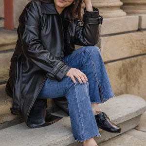 Pu&ograve; includere: Una persona indossa un trench coat in pelle nera, jeans blu e scarpe nere stringate. I jeans hanno uno spacco alla caviglia. La persona &egrave; seduta su gradini di pietra.