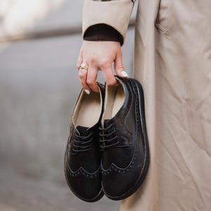 Puede incluir: Un par de zapatos Oxford de cuero negro con perforaciones decorativas y cierres de cordones. Los zapatos son sostenidos por una persona que lleva una gabardina beige.