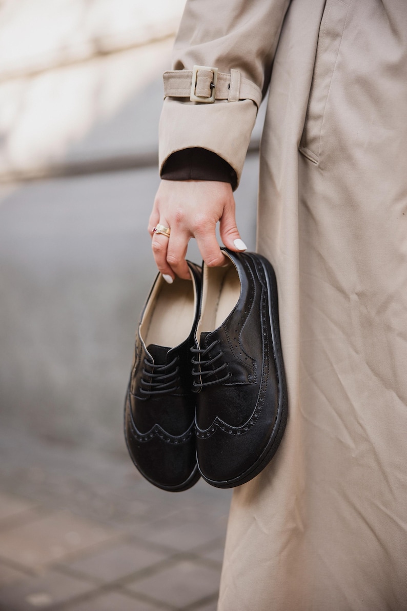 Puede incluir: Un par de zapatos Oxford de cuero negro con perforaciones decorativas y cierres de cordones. Los zapatos son sostenidos por una persona que lleva una gabardina beige.
