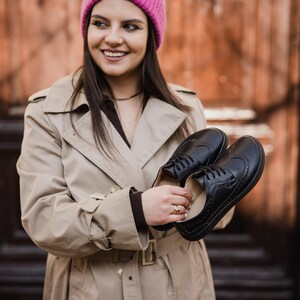 Puede incluir: Un par de zapatos brogue de cuero negro con perforaciones decorativas. Los zapatos son sostenidos por una persona que lleva una gabardina beige y un gorro de punto rosa. Dise&ntilde;o cl&aacute;sico.