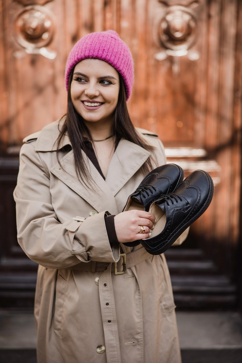 Puede incluir: Un par de zapatos brogue de cuero negro con perforaciones decorativas. Los zapatos son sostenidos por una persona que lleva una gabardina beige y un gorro de punto rosa. Dise&ntilde;o cl&aacute;sico.