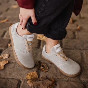 May include: Light gray suede sneakers with brown soles and beige laces. The sneakers have a minimalist design with a white accent on the heel and a logo on the tongue. The shoes are shown on a brick surface.