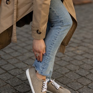 May include: A pair of light brown canvas trainers with white laces and a white sole. The trainers are being worn by a person wearing blue jeans and a beige trench coat.