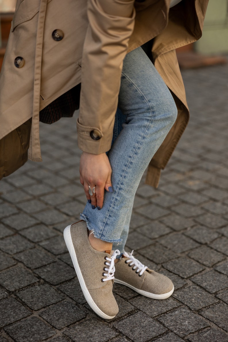 May include: A pair of light brown canvas trainers with white laces and a white sole. The trainers are being worn by a person wearing blue jeans and a beige trench coat.