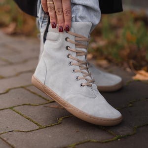 Könnte beinhalten: Hellgraue Wildleder-Ankle Boots mit beigefarbenen Schnürsenkeln und Sohlen. Die Stiefel haben ein minimalistisches Design mit runder Spitze und einer hoch geschnittenen Silhouette. Die Stiefel sind geschnürt und scheinen aus weichem Wildleder zu sein.