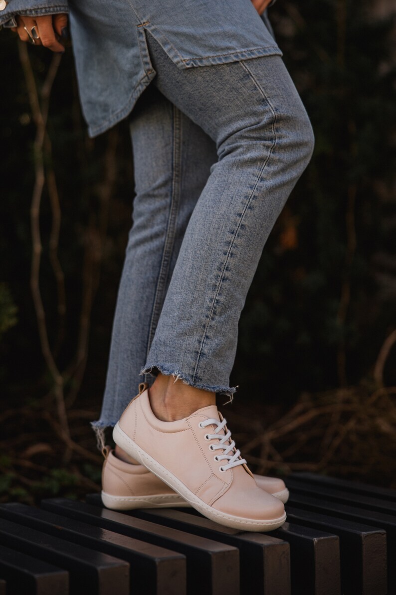 May include: A pair of light pink leather sneakers with white laces. The sneakers are on a black and brown striped surface.