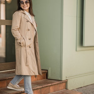 May include: A woman wearing a beige trench coat, blue jeans, and beige trainers stands on a set of brown steps in front of a building with a green wall.