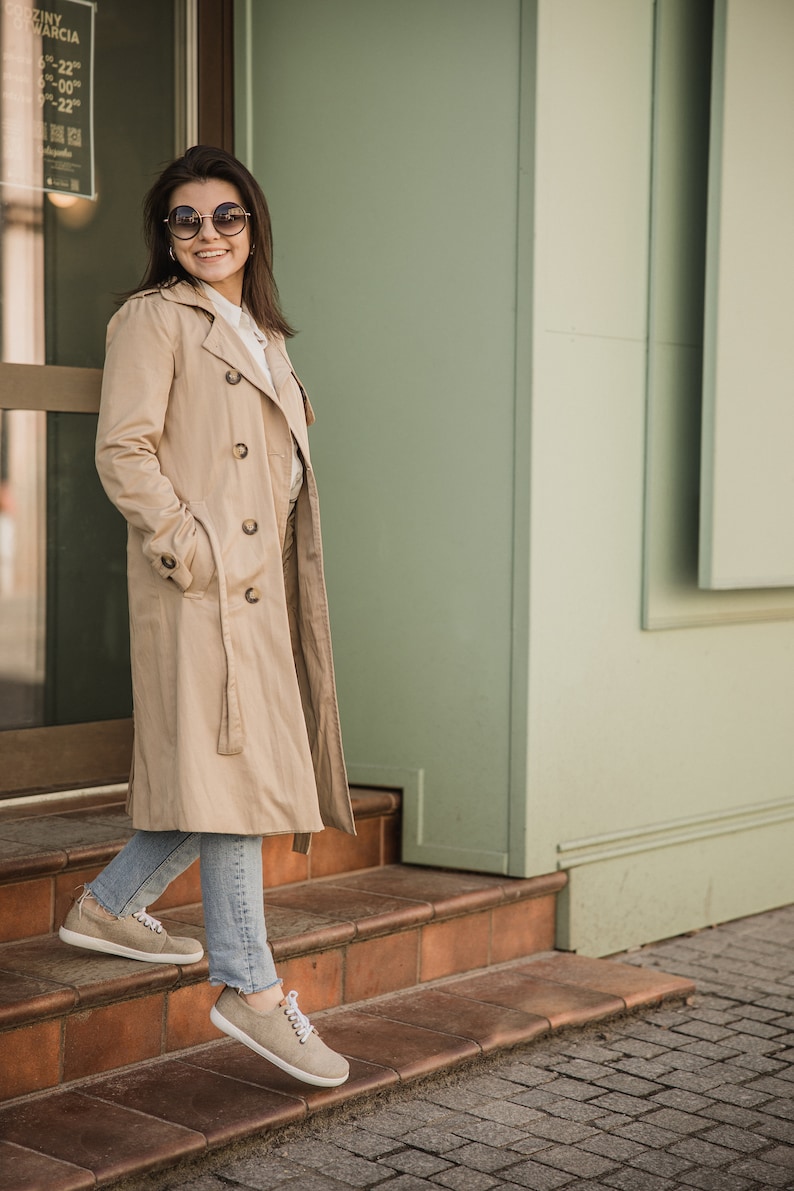 May include: A woman wearing a beige trench coat, blue jeans, and beige trainers stands on a set of brown steps in front of a building with a green wall.