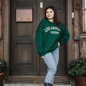 Op de afbeelding: Een vrouw draagt een groen sweatshirt met de tekst "Los Angeles, Californi&euml;" op de voorkant, een blauwe jeans en zwarte veterschoenen. Ze staat voor een houten deur.