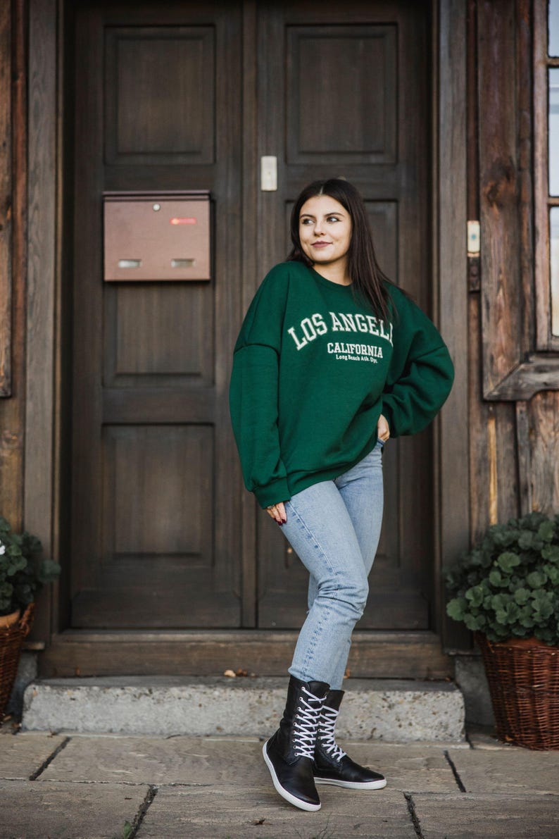 Op de afbeelding: Een vrouw draagt een groen sweatshirt met de tekst "Los Angeles, Californi&euml;" op de voorkant, een blauwe jeans en zwarte veterschoenen. Ze staat voor een houten deur.