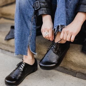 Pu&ograve; includere: Scarpe brogue in pelle nera con finitura lucida. Le scarpe presentano un classico design a punta di ala con perforazioni decorative e chiusura con lacci. L'immagine mostra le scarpe che vengono allacciate, suggerendo che vengono indossate.
