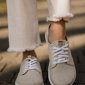 May include: A pair of beige canvas sneakers with white laces. The sneakers have a white rubber sole and are worn by a person wearing white pants.
